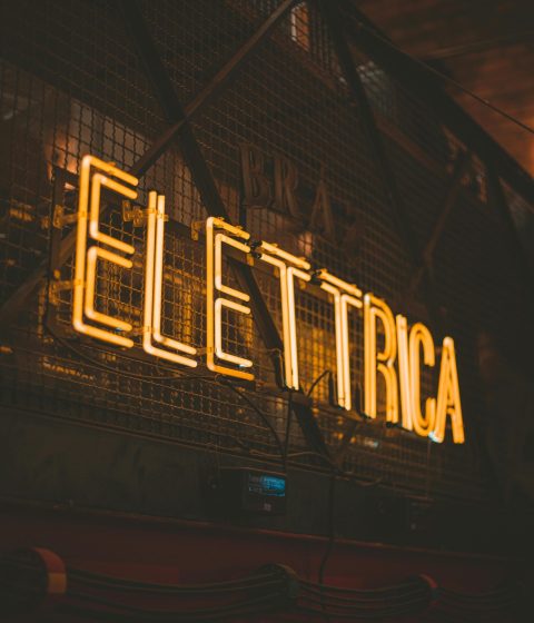 Moody urban photo of an illuminated 'Elettrica' neon sign in warm tones.